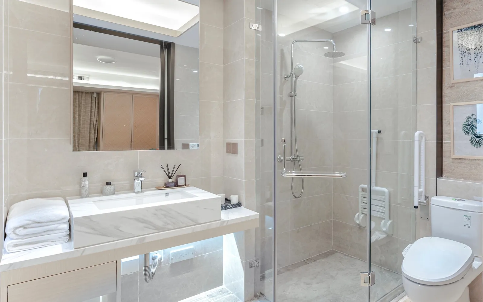 Salle de bain claire aux carreaux beige : douche d'angle vitrée de plain-pied équipée d'un siège rabattable blanc, d'une barre d'appui verticale et d'une barre horizontale sous la colonne de douche, meuble vasque et WC à côté
