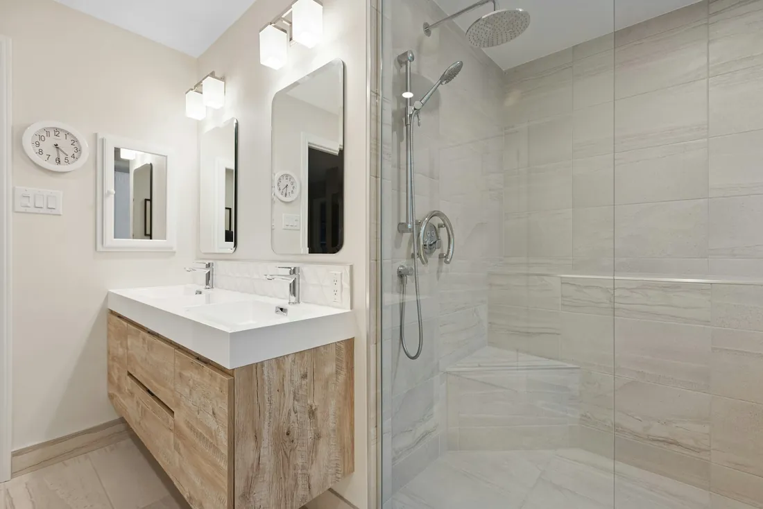 Douche à l'italienne derrière une paroi vitrée, avec une barre d'appui arrondie près du mitigeur, une banquette carrelée et une douchette sur barre ; meuble vasque en bois clair à gauche (Bucy-le-Long, Aisne)