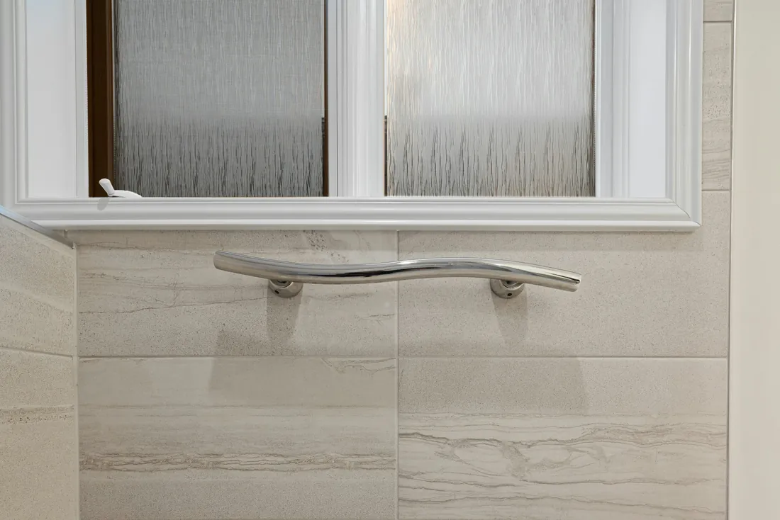 Barre d'appui chromée galbée fixée sur le carrelage beige d'une douche, sous une fenêtre en verre dépoli (Beuvry-la-Forêt, Nord)