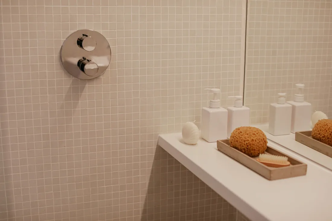 Mitigeur thermostatique encastré à deux commandes rondes sur une mosaïque beige, à côté d'une tablette avec flacons et éponge naturelle (Plounéour-Brignogan-plages, Finistère)