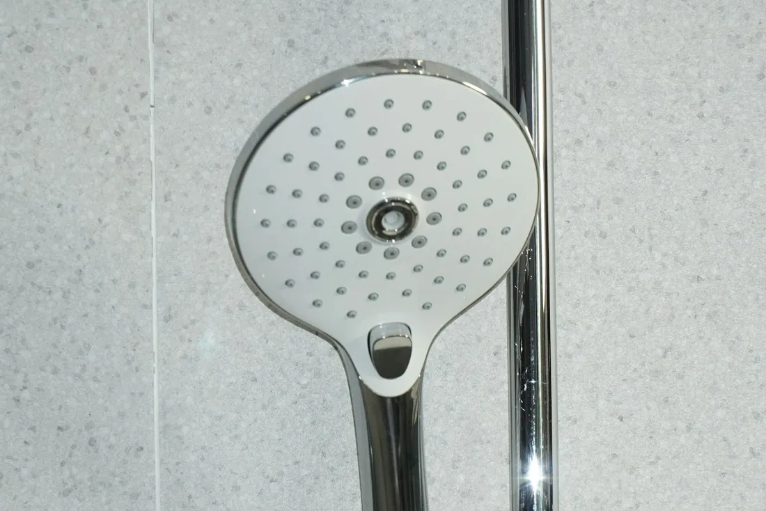 Douchette à main blanche et chromée à jets larges, accrochée à sa barre de douche devant un mur gris clair (Chenoise-Cucharmoy, Seine-et-Marne)