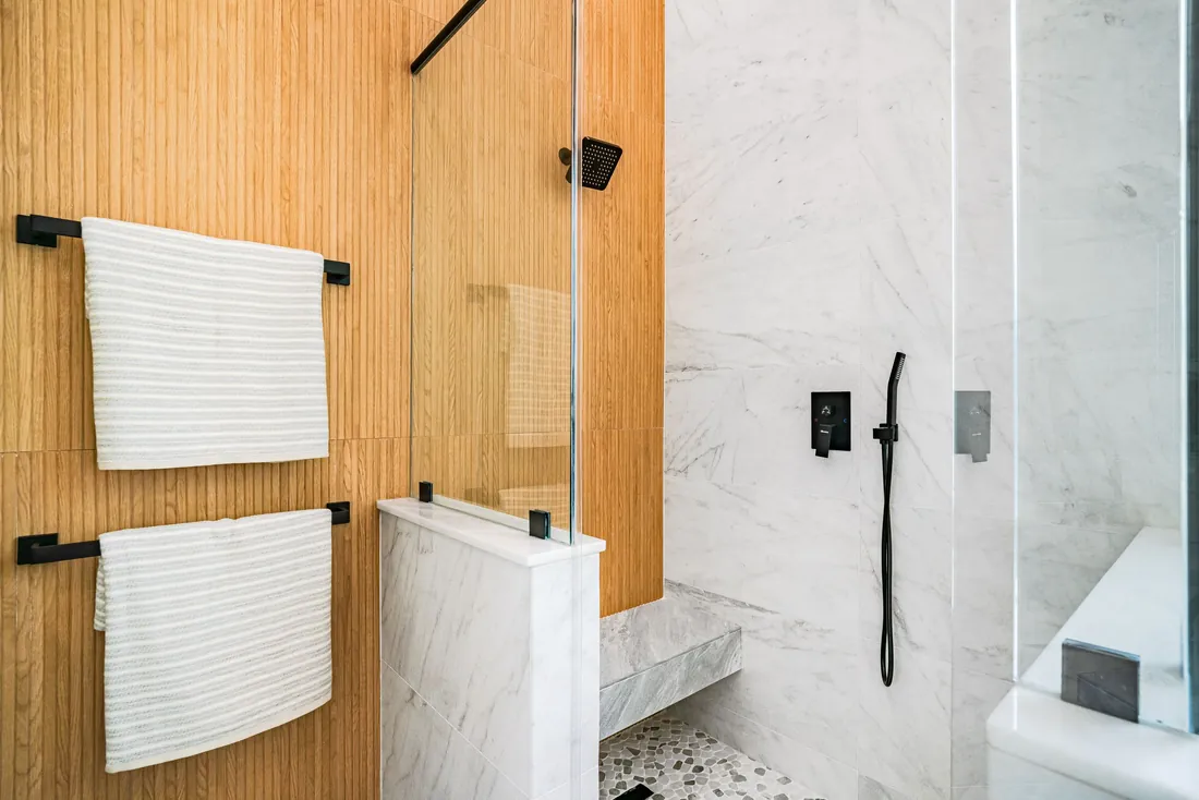 Douche en marbre blanc avec banc maçonné, douchette noire sur flexible et paroi vitrée, porte-serviettes noirs sur un mur à lattes de bois (Périgny, Val-de-Marne)
