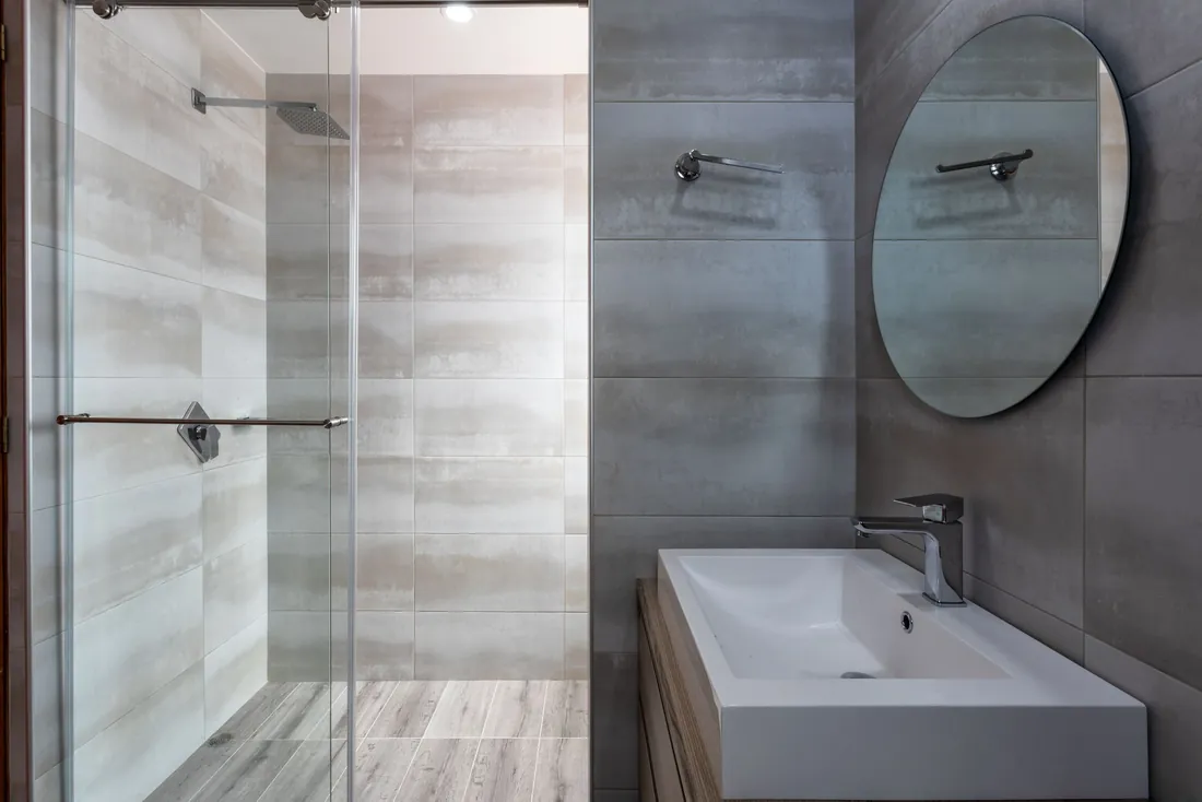 Douche vitrée au carrelage gris clair avec barre horizontale sur la porte, à côté d'un lavabo blanc rectangulaire et d'un miroir rond sur un mur gris anthracite (Naves, Corrèze)