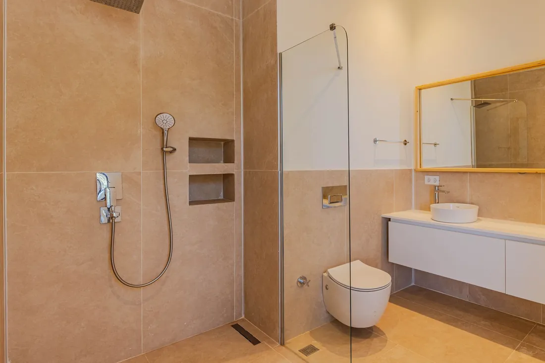 Douche de plain-pied au carrelage beige avec paroi vitrée fixe, mitigeur et douchette sur flexible, deux niches murales et caniveau au sol ; WC suspendu et meuble vasque blanc au fond (Heuringhem, Pas-de-Calais)
