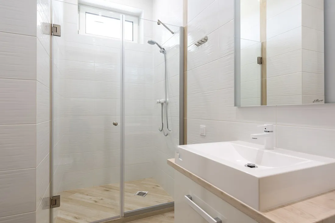 Douche fermée par deux portes vitrées, faïence blanche texturée, sol effet parquet clair et fenêtre en haut du mur ; grand lavabo blanc sur plan bois au premier plan (Barbizon, Seine-et-Marne)
