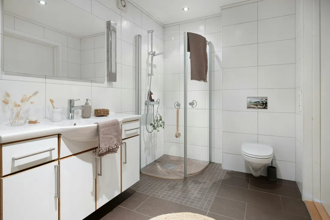 Salle de bain toute blanche avec douche quart de cercle sans seuil à parois vitrées, douchette sur barre et brosse suspendue, meuble vasque blanc et WC suspendu sur un carrelage brun (Craon, Mayenne)