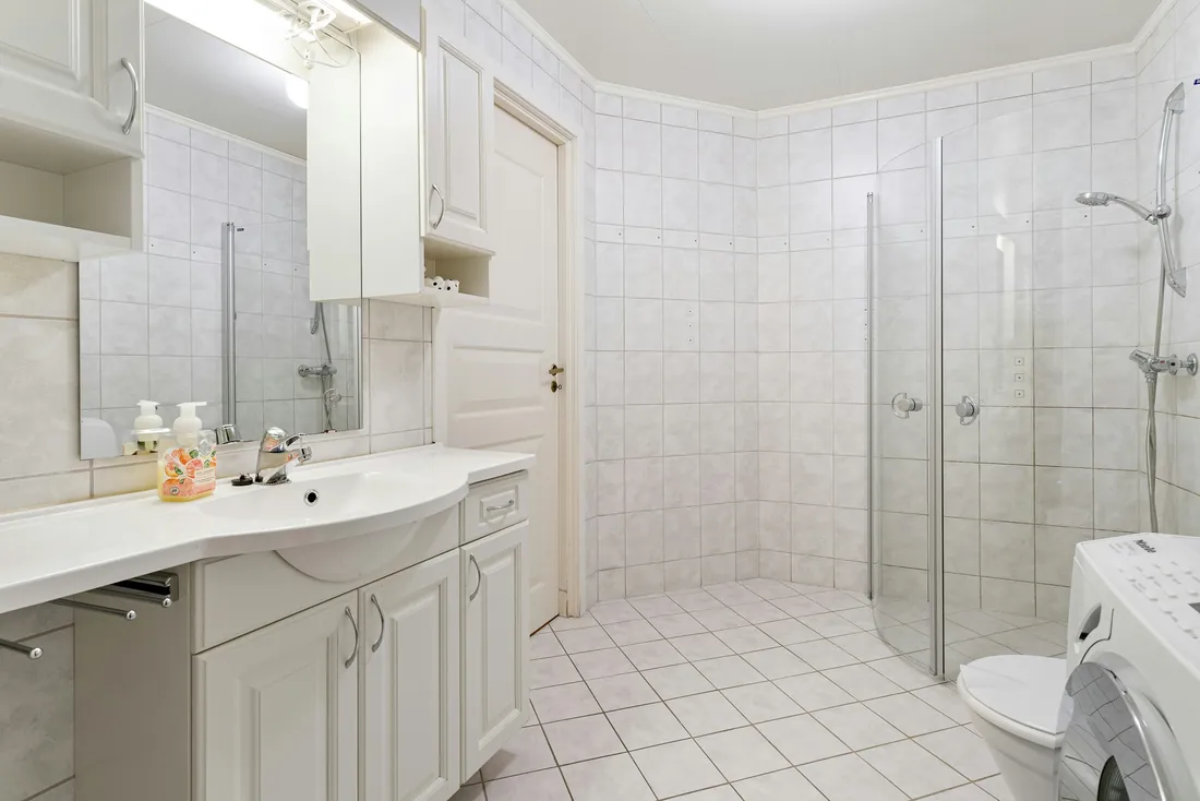 Salle de bain carrelée de blanc avec douche d'angle de plain-pied à parois vitrées repliables, meuble lavabo à portes moulurées et lave-linge à côté du WC (Thise, Doubs)