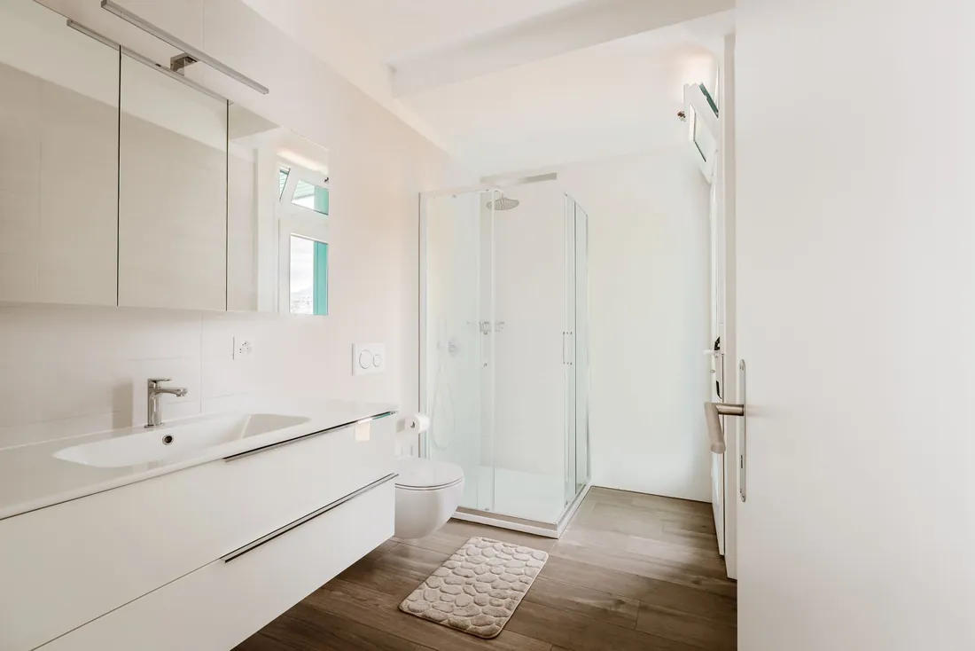 Salle de bain blanche épurée : cabine de douche d'angle vitrée à receveur bas, long plan vasque suspendu, armoire de toilette et sol effet parquet (Propriano, Corse-du-Sud)