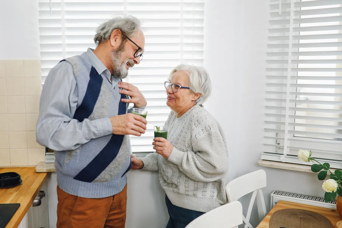 Couple de seniors souriants qui trinquent avec un jus vert dans une cuisine claire aux stores blancs
