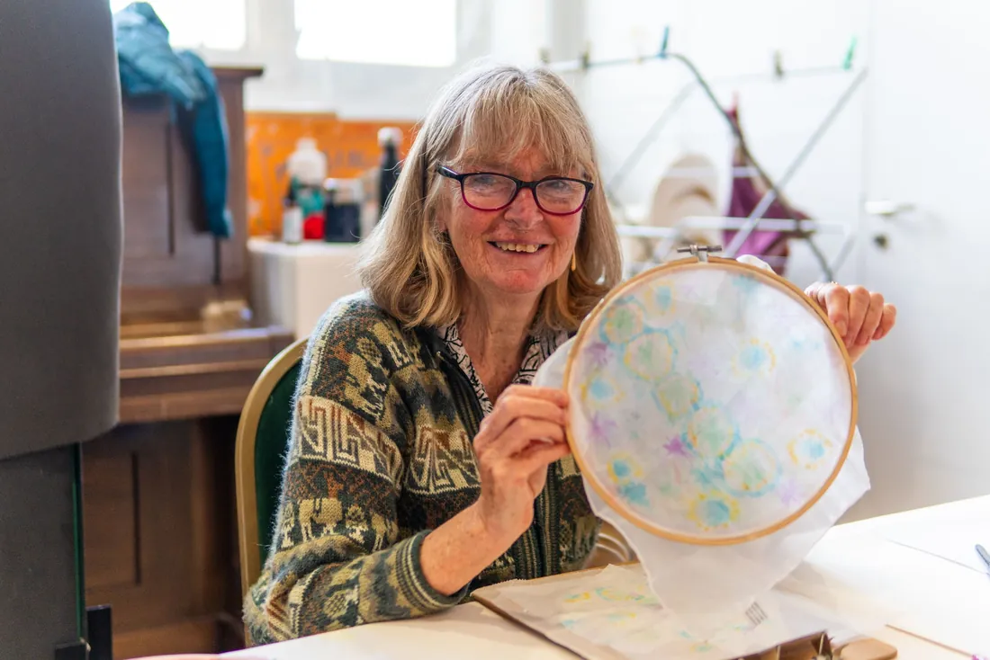Femme sénior à lunettes, souriante, montrant sa broderie sur tambour, assise à une table chez elle