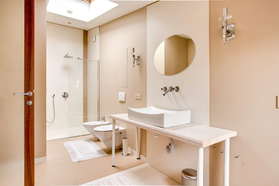 Salle de bain beige et lumineuse : douche à l'italienne au fond derrière une paroi vitrée, WC et bidet suspendus, plan vasque sur pieds blancs (Cabestany, Pyrénées-Orientales)